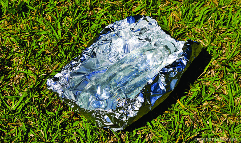 A photo of a clear bottle filled with water resting on a sheet of aluminum foil in the sun as a water purification method.